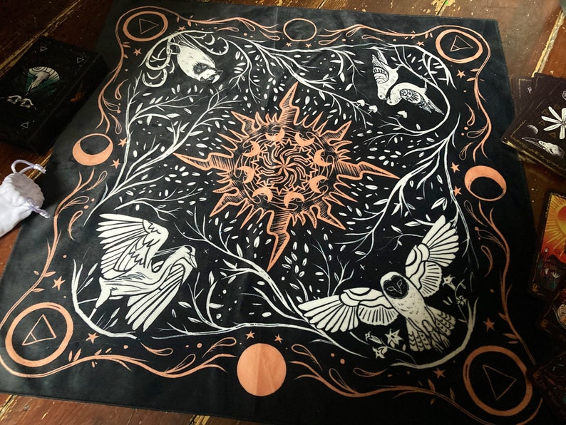 May include: Black fabric tapestry with a white and peach design. The design features a sun with rays, two birds, and a crescent moon. The tapestry has a border of peach and white.