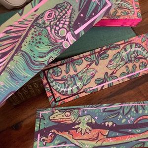 May include: Set of three bookmarks featuring colourful illustrations of lizards on a green background with pink borders. The bookmarks are printed on a textured paper with a metallic sheen.