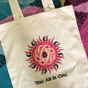 May include: A white tote bag with a pink and black sun design and the text "The All is One".