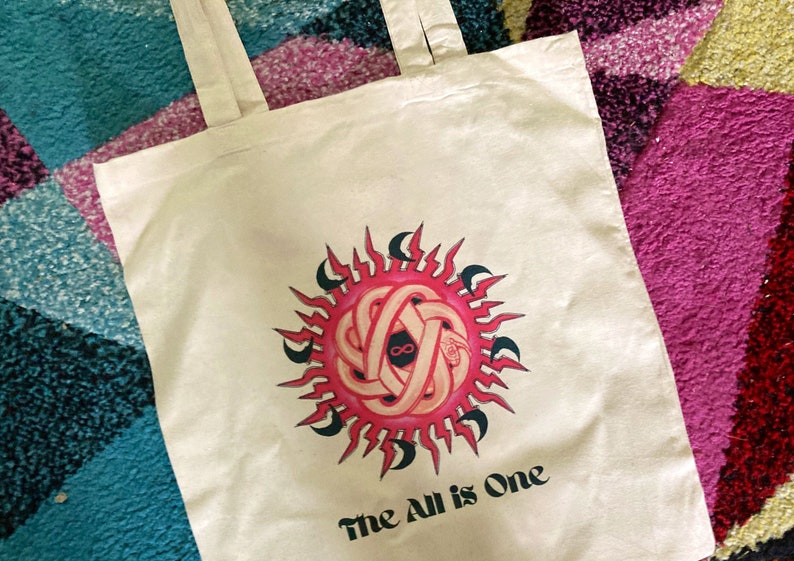 May include: A white tote bag with a pink and black sun design and the text "The All is One".