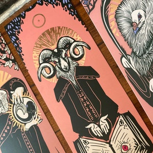 May include: A pink bookmark featuring a black and white illustration of a ram-headed figure in a black robe, holding an open book with two red eyes. The book rests on a table with a gold rim. The background is a pink and purple floral design.