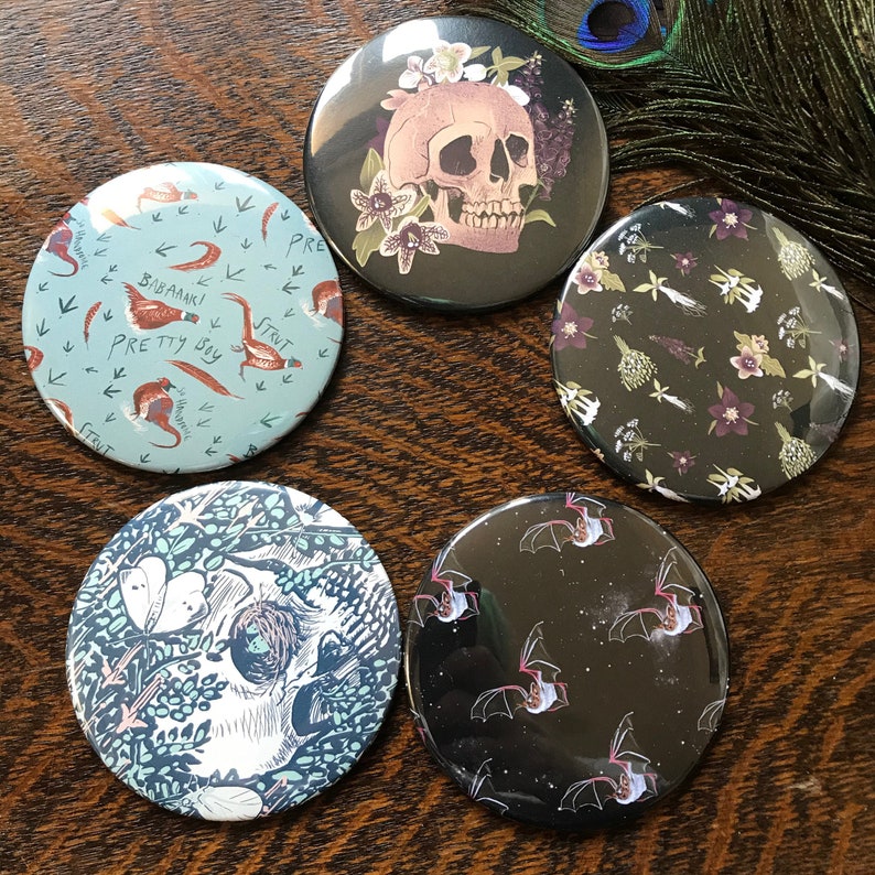May include: Set of five round, 2.25 inch diameter,  mirror-like buttons with various designs. One button features a skull with flowers, another has a black background with white bats, another has a black background with white flowers, another has a blue background with birds and the text "Pretty Boy Strut", and the last one has a blue background with a skull and leaves.