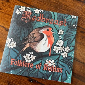 May include: A teal blue book cover with a red robin bird illustration and white flowers. The title of the book is "Redbreast Folklore of Robins".