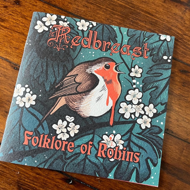 May include: A teal blue book cover with a red robin bird illustration and white flowers. The title of the book is "Redbreast Folklore of Robins".