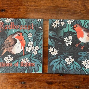 May include: Two cards with a robin illustration and the text "The Redbreast Folklore of Robins". The cards have a dark green background with white flowers and a robin with a red breast.