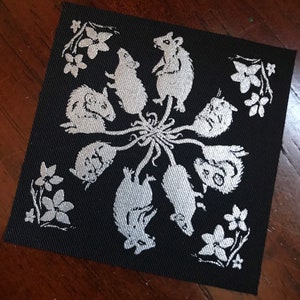 May include: A black fabric patch with a white design of eight mice in a circle around a central flower. The mice are facing outward and have long tails. The design is reminiscent of a mandala.