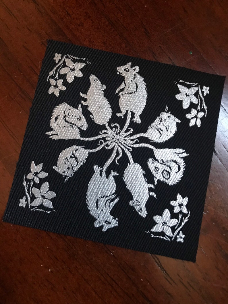May include: A black fabric patch with a white design of eight mice in a circle around a central flower. The mice are facing outward and have long tails. The design is reminiscent of a mandala.