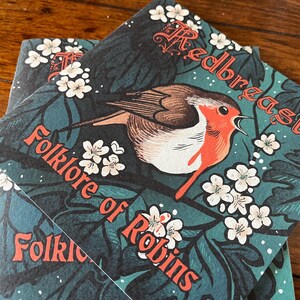 May include: Two book covers with a green background and white flowers. The top cover features a red robin with the text "Redbreast Folklore of Robins". The bottom cover has the text "Folklore of Robins" in red.