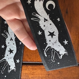 May include: Black rectangular patch with a silver fox design, stars, and a crescent moon. The fox is depicted in a stylized, celestial pose, with a star at its center. The patch is likely for clothing or accessories.