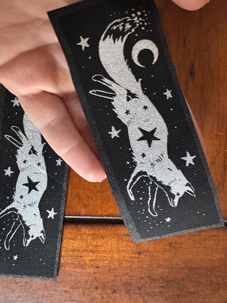May include: Black rectangular patch with a silver fox design, stars, and a crescent moon. The fox is depicted in a stylized, celestial pose, with a star at its center. The patch is likely for clothing or accessories.