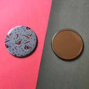 May include: A round, blue compact mirror with a design of brown pheasants and the text "Strut Pretty Boy" on a white background. The mirror is on a pink and gray surface.