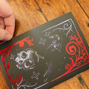 May include: A black card with a red and silver design. The design features a skull, a raven, and floral elements.