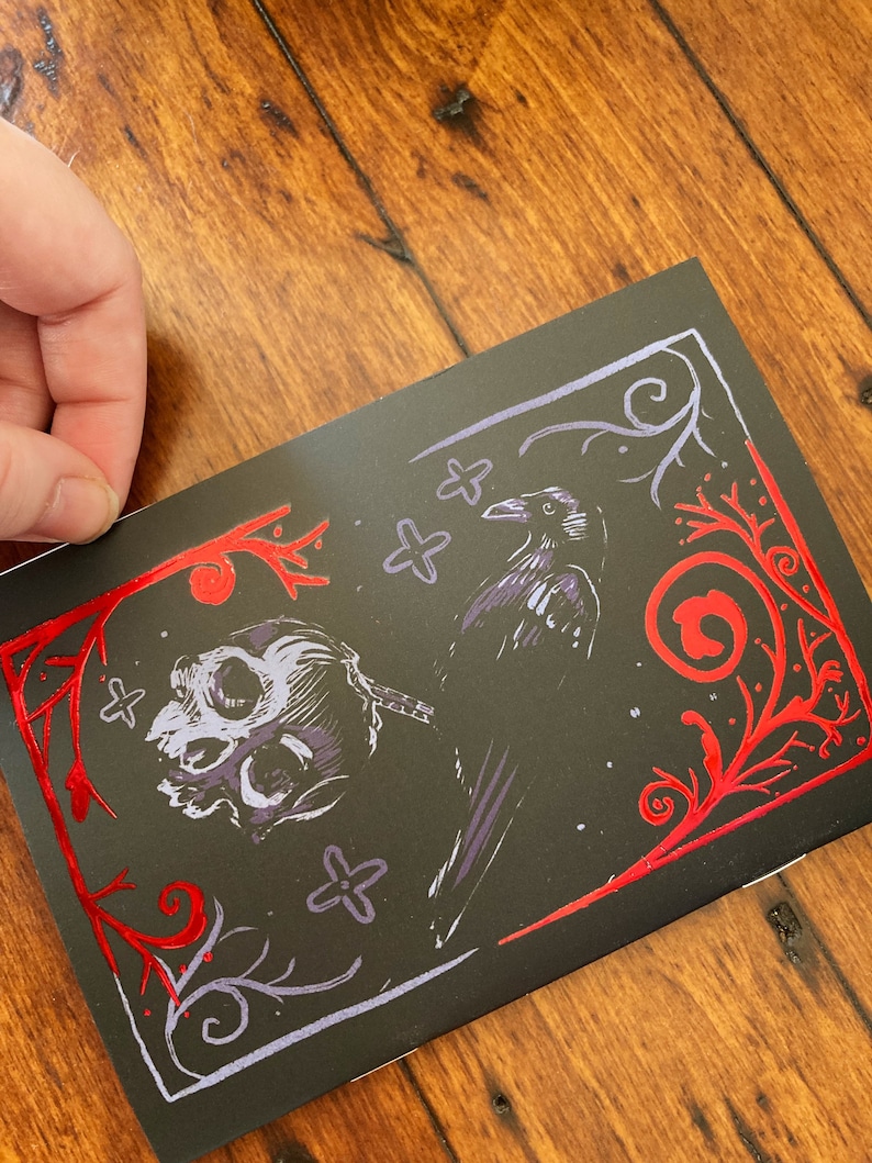 May include: A black card with a red and silver design. The design features a skull, a raven, and floral elements.