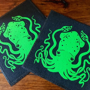 May include: Two black fabric patches with a green squid design. The squid is outlined in black and has a spotted pattern. The squid has two large eyes and eight tentacles.