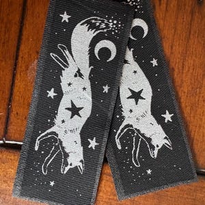 May include: Two black rectangular patches with a silver fox design, stars, and crescent moons. The fox is upside down with a star on its belly. The patches are on a wooden surface.