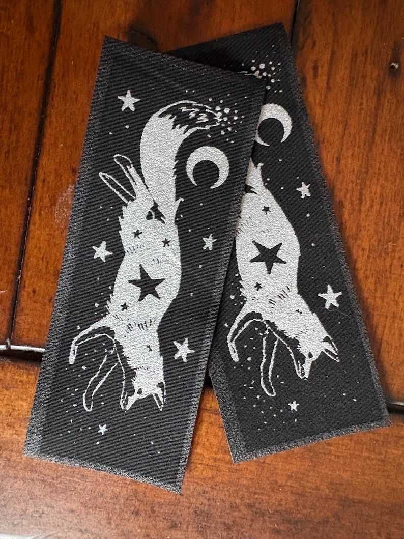 May include: Two black rectangular patches with a silver fox design, stars, and crescent moons. The fox is upside down with a star on its belly. The patches are on a wooden surface.