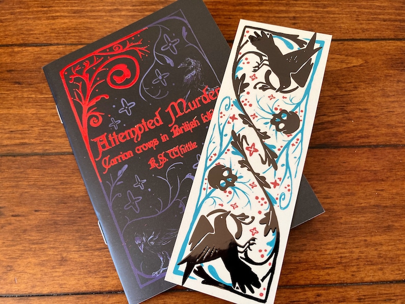 May include: A black book with a red title that reads "Attempted Murder: Carrion Crows in British Birds" by R.S. Whittle. The book is accompanied by a bookmark with a black and white illustration of birds and skulls on a white background with blue and red accents.