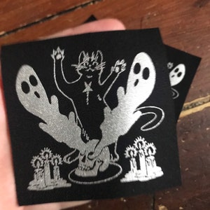 May include: Two black square patches with a silver graphic design. The design features a cat with a star on its chest, a skull, ghosts, and candles. The patches are likely for clothing or accessories.