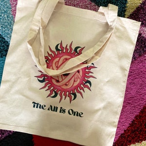 May include: A white canvas tote bag with a pink and black sun design and the text "The All is One".