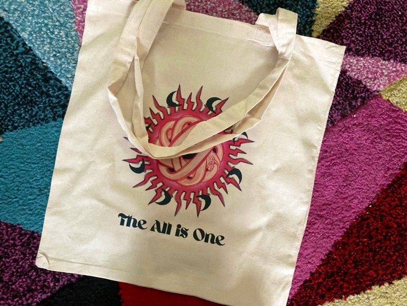 May include: A white canvas tote bag with a pink and black sun design and the text "The All is One".