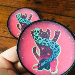 May include: Two pink and black embroidered iron-on patches featuring a black cat with a blue and white patterned scarf. The cat is in a playful pose with its paw raised.