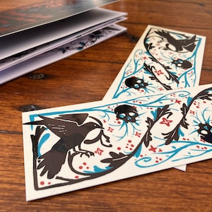 May include: Two black and white bookmarks featuring a black bird, skulls, and floral designs on a teal background. The bookmarks have red accents.
