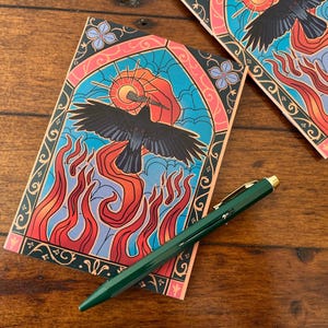 May include: A black crow with red flames behind it, flying through a stained glass window with a blue and orange sky. The window is framed in black and has a pink and green floral design.