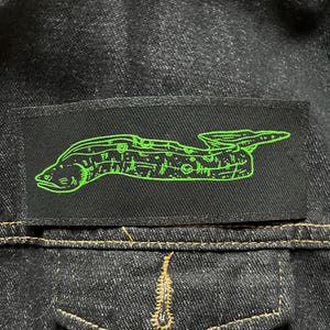 May include: A black patch featuring a green embroidered image of a moray eel. The eel is depicted in a stylised, almost cartoonish way, with a large head and a long, slender body.