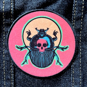 May include: A round, pink and green patch with a black and blue beetle design. The beetle has a skull for a head and is surrounded by a peach-colored halo.