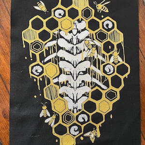 May include: Black artwork with a gold honeycomb design and a white spine-like structure. Bees and larvae are scattered around the honeycomb. The artwork has a hand-drawn aesthetic, with a dripping effect.