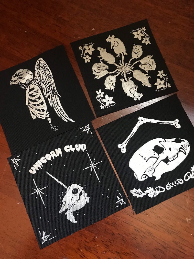May include: Four black fabric patches with silver-colored designs. The designs include a skeleton with wings, a circle of rats, a skull with bones, and a unicorn skull with the text "Unicorn Club".