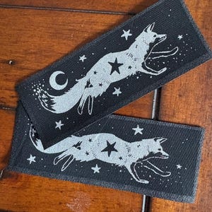 May include: Two rectangular black patches with a silver fox design leaping among stars and a crescent moon. The patches are made of a textured fabric, and the fox and celestial elements are detailed.
