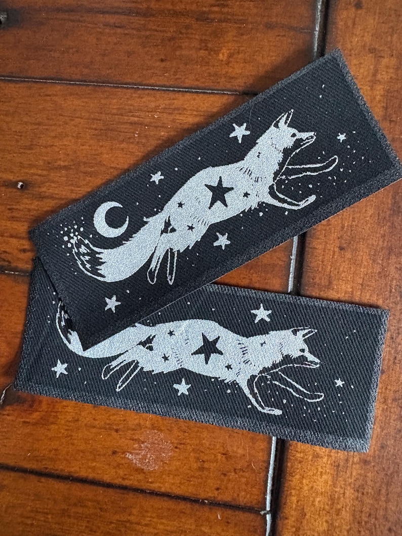 May include: Two rectangular black patches with a silver fox design leaping among stars and a crescent moon. The patches are made of a textured fabric, and the fox and celestial elements are detailed.