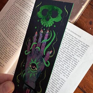 May include: A black bookmark with a green skull and a hand with an eye design. The hand has long, pointed fingernails and is wrapped in green vines. The bookmark is set against the backdrop of an open book.