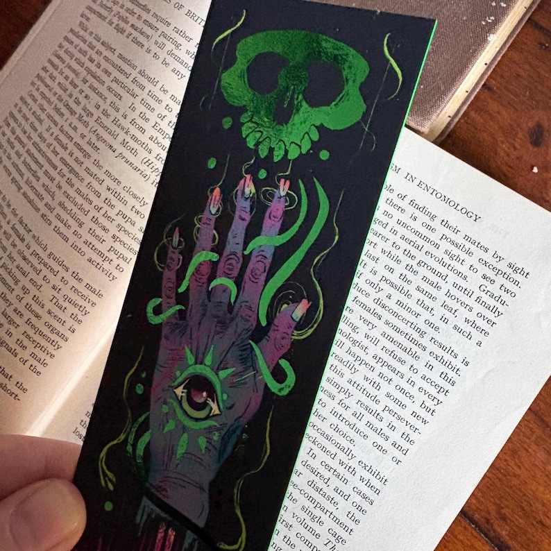 May include: A black bookmark with a green skull and a hand with an eye design. The hand has long, pointed fingernails and is wrapped in green vines. The bookmark is set against the backdrop of an open book.