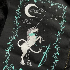 Op de afbeelding: Een zwarte stoffen tas met een wit en turquoise ontwerp van een eenhoorn met een zwaard en een halve maan. Het ontwerp is omgeven door een bladrankenpatroon.