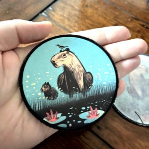 May include: A round, blue and black patch with a capybara, a small rodent, and a bird. The capybara is brown and has a white face. The rodent is brown and white. The bird is black. The patch has a black border and is surrounded by a white background.