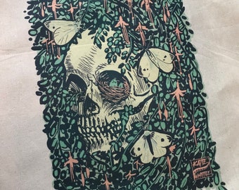 May include: A black and white illustration of a skull surrounded by green foliage and butterflies. The skull has a detailed design with a bird's nest in the eye socket. The text "Katie Whittle" is in the lower right corner.