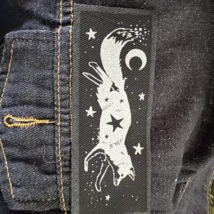 May include: A black rectangular patch featuring a white fox design with a crescent moon and stars. The fox is depicted upside down, with a large star in the center of its body. The patch is sewn onto a dark denim jacket.