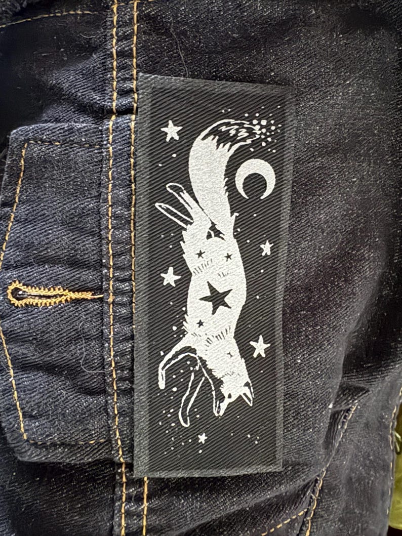 May include: A black rectangular patch featuring a white fox design with a crescent moon and stars. The fox is depicted upside down, with a large star in the center of its body. The patch is sewn onto a dark denim jacket.