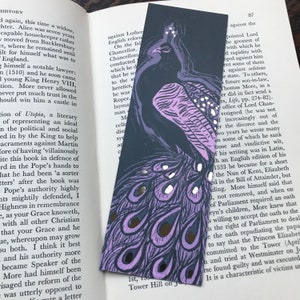 Może przedstawiać: Zakładka z ilustracją purpurowego i czarnego pawia z złotymi akcentami. Zakładka jest ciemnoszara z ilustracją purpurowego i czarnego pawia z złotymi akcentami. Tekst "Tower Hill on July 1st, 1535" jest nadrukowany na dole zakładki.