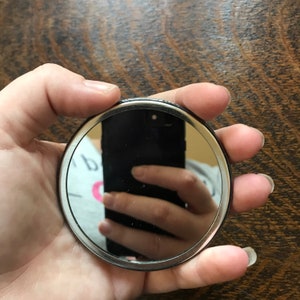 May include: A silver-colored compact mirror with a black rim. The mirror is reflecting a hand holding a black smartphone.