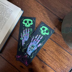 May include: Two black bookmarks with a spooky design. Each bookmark features a green skull above a purple hand with an eye in the palm. The hand has long, pointed nails and is surrounded by green, snake-like lines.
