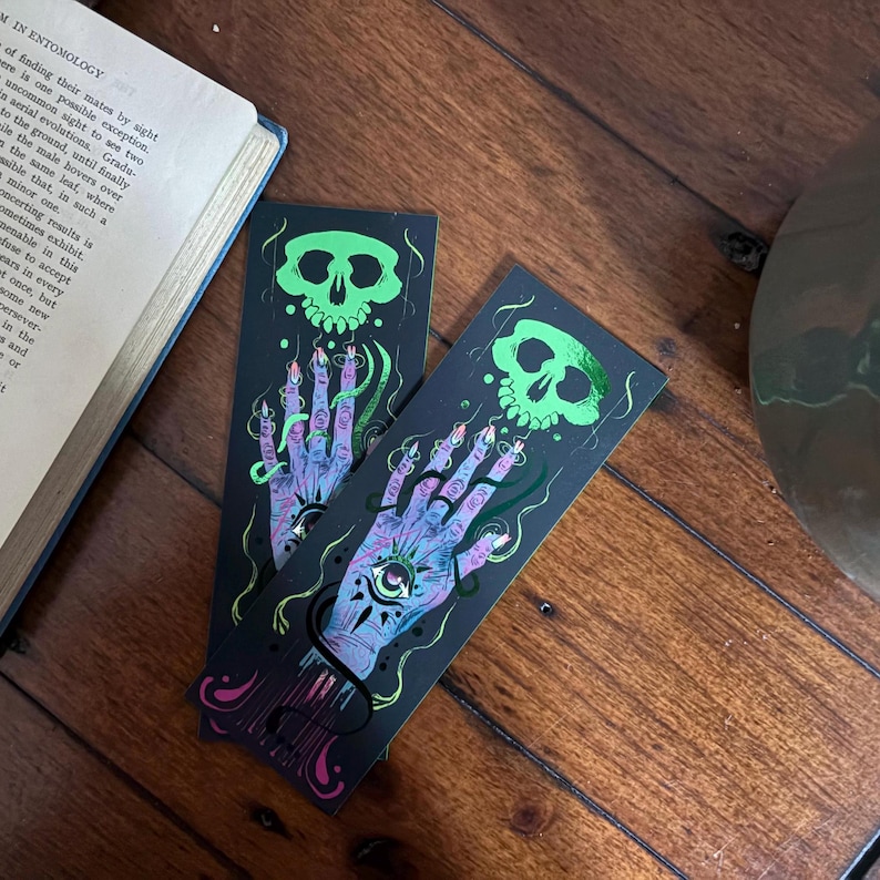 May include: Two black bookmarks with a spooky design. Each bookmark features a green skull above a purple hand with an eye in the palm. The hand has long, pointed nails and is surrounded by green, snake-like lines.