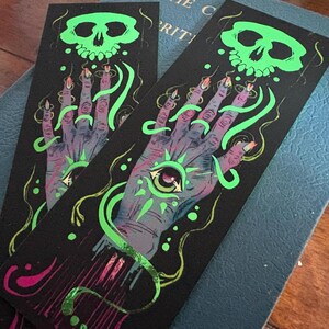 May include: Two black bookmarks with a spooky design. Each bookmark features a purple hand with a glowing green eye, a green skull, and green, glowing tendrils. The nails are painted with orange and pink. The design is printed on a black background.
