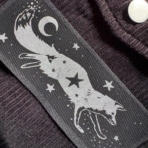 May include: A rectangular black patch with a silver fox design, stars, and a crescent moon. The fox is leaping, with a star on its body. The patch is on a dark brown, ribbed fabric.