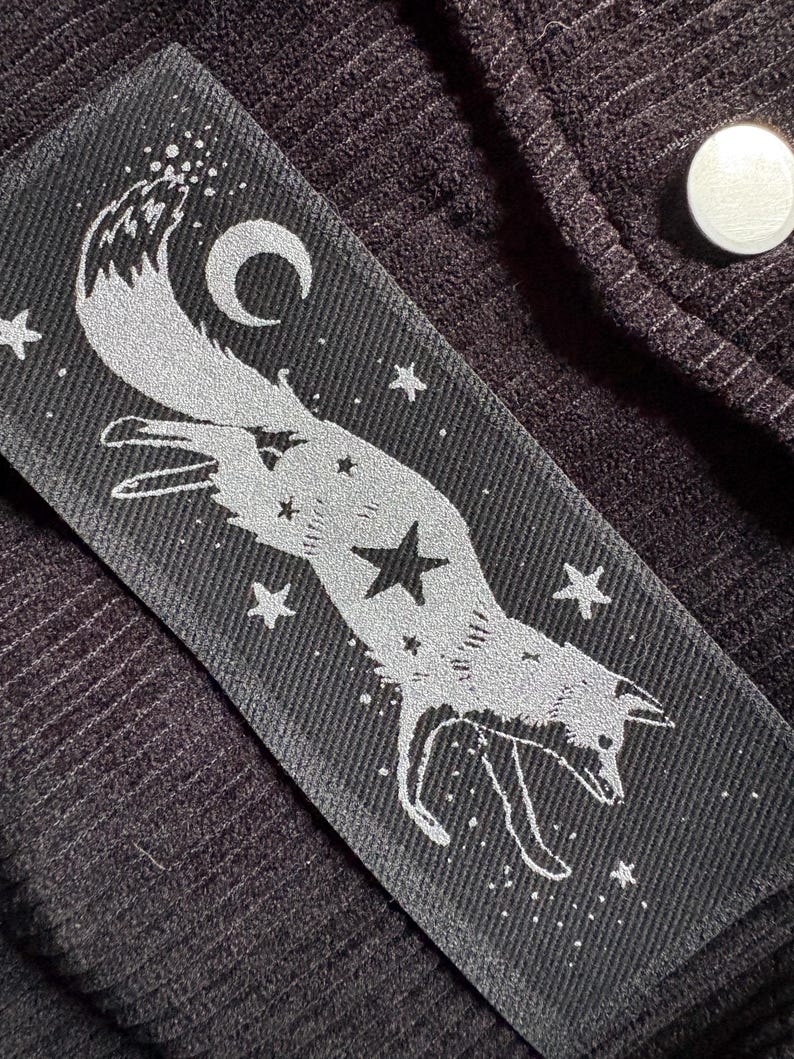 May include: A rectangular black patch with a silver fox design, stars, and a crescent moon. The fox is leaping, with a star on its body. The patch is on a dark brown, ribbed fabric.