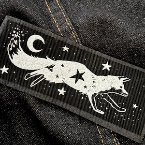 May include: A black rectangular patch with a white fox design, a crescent moon, and stars. The fox is depicted running, with a constellation theme. The patch is likely for clothing or accessories, and is made of fabric.