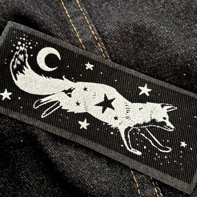 May include: Black rectangular patch featuring a white fox silhouette with a crescent moon and stars. The fox is running, and the design includes a constellation theme. The patch is likely for clothing or accessories.