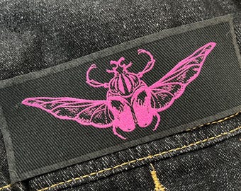 May include: A black rectangular patch featuring a pink beetle design with outstretched wings. The patch is sewn onto dark denim fabric with yellow stitching. The beetle is detailed with intricate lines.